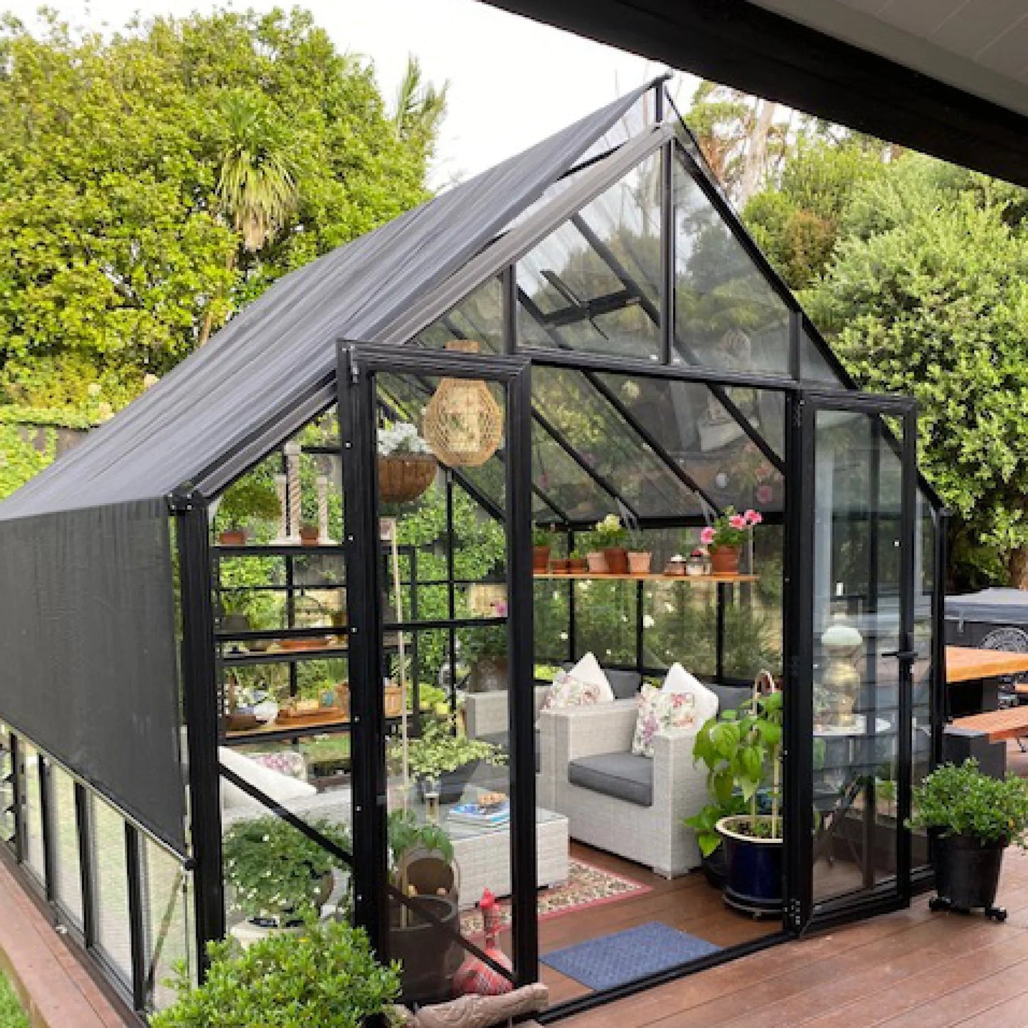 Modern black-framed garden greenhouse with glass panels, shade cover, and cozy seating area